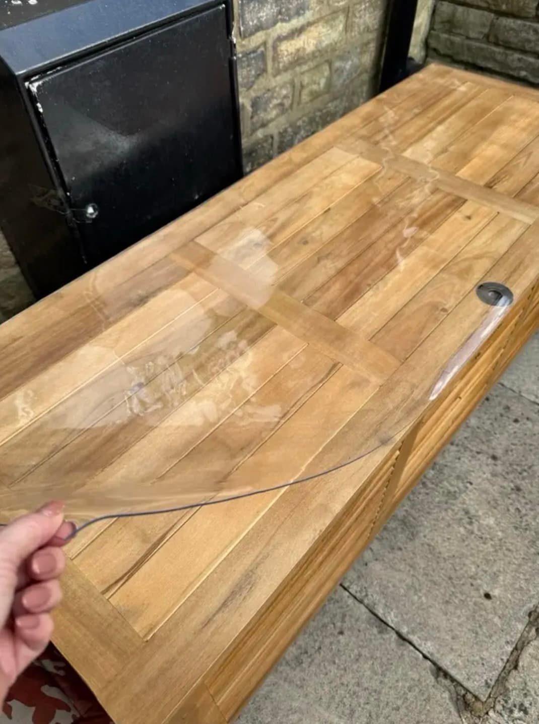 Banco de jardín en listones de teca bajo película transparente; una mano sostiene la esquina